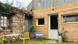 Un rústico edificio de madera con un muro de piedra, una puerta y una pequeña ventana. En primer plano, hay una mesa y dos sillas.