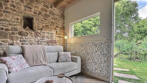 Un muro de piedra, un sofá gris con cojines estampados y una ventana con vistas a la vegetación.