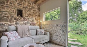 Eine Steinsäule, ein graues Sofa mit floralen Kissen und ein Fenster mit Blick auf Grünflächen.