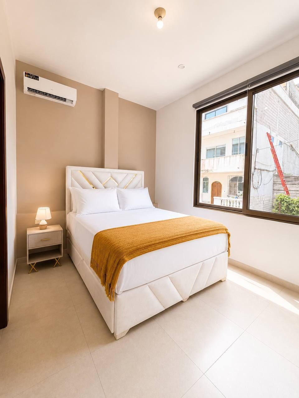 Quarto com cama, mesa de cabeceira, ar condicionado, janela com vista para edifícios e luminária no teto.