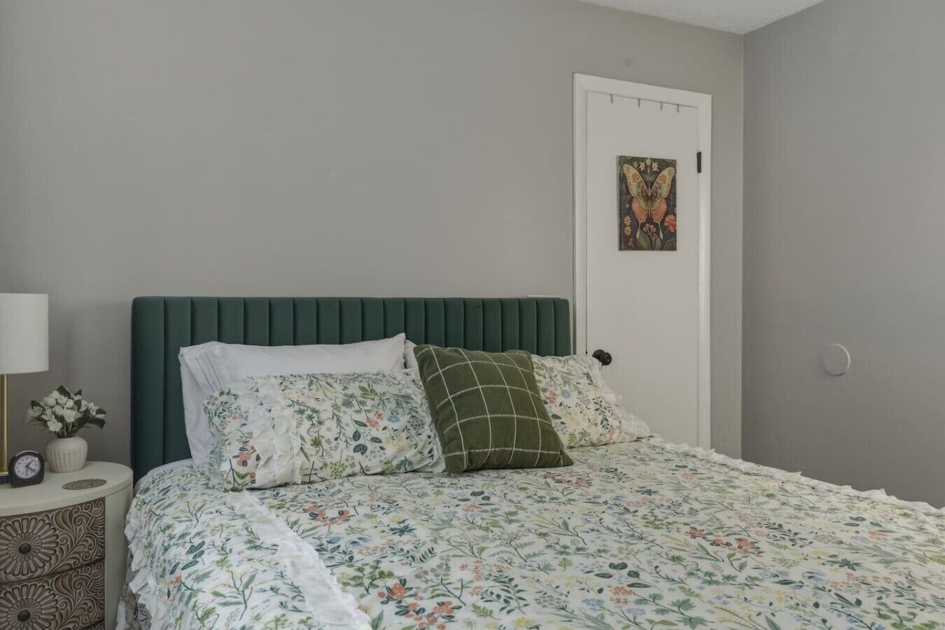 Une chambre à coucher avec une tête de lit recouverte d’un tissu vert, une couvre-lit à motifs floraux et un seul tableau accroché au mur.