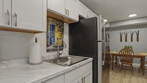 Een keuken met witte kasten, een zwarte koelkast, een marmeren aanrecht, een spoelbak en een eethoek met houten stoelen en een tafel.