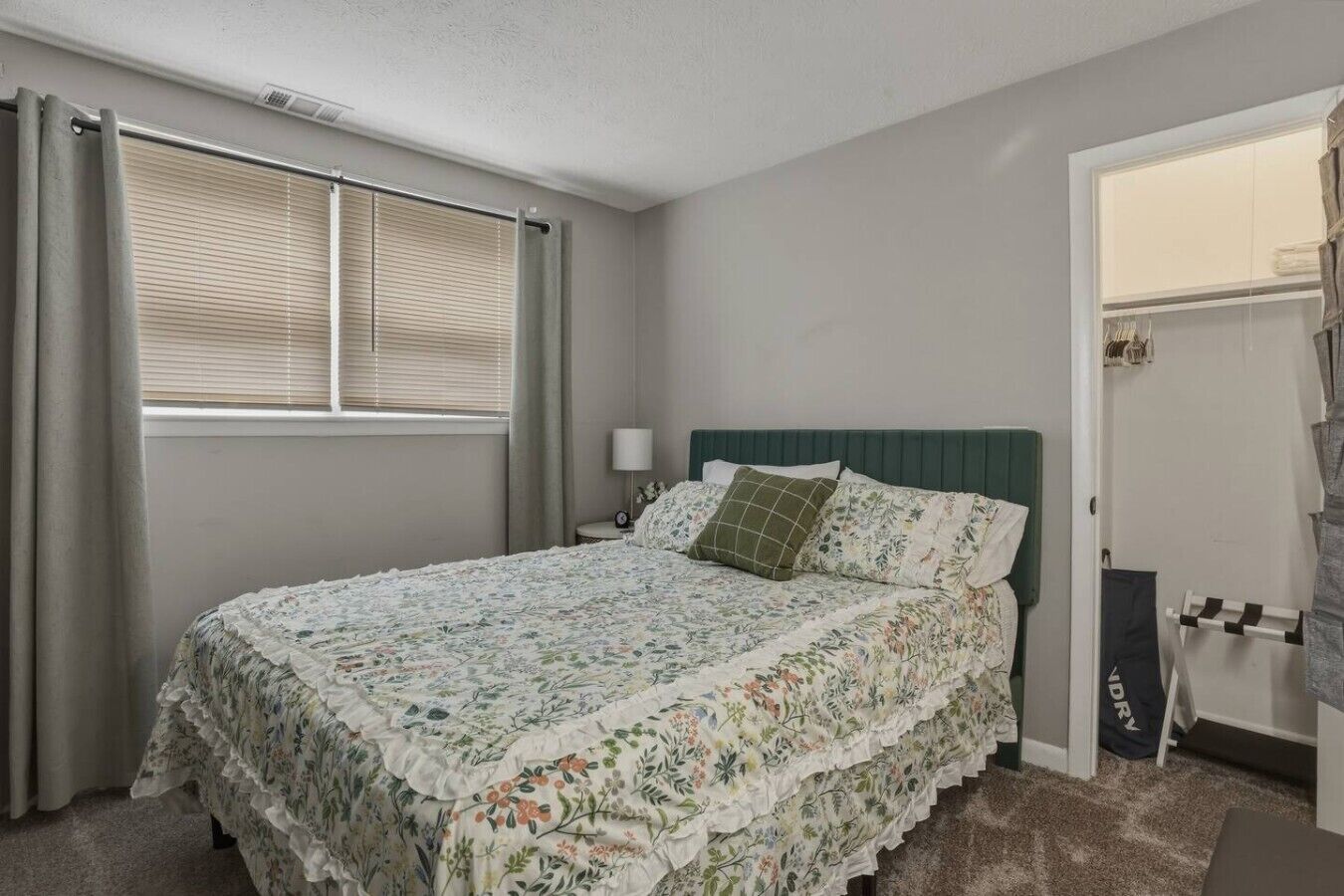 A bedroom with a bed, a nightstand, a lamp, a window with blinds, and a door leading to a closet.