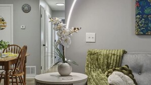 Een woonkamer met een grijze bank, een rond bijzettafeltje met een vaas witte bloemen, een groene plaid en een decoratief wandobject.