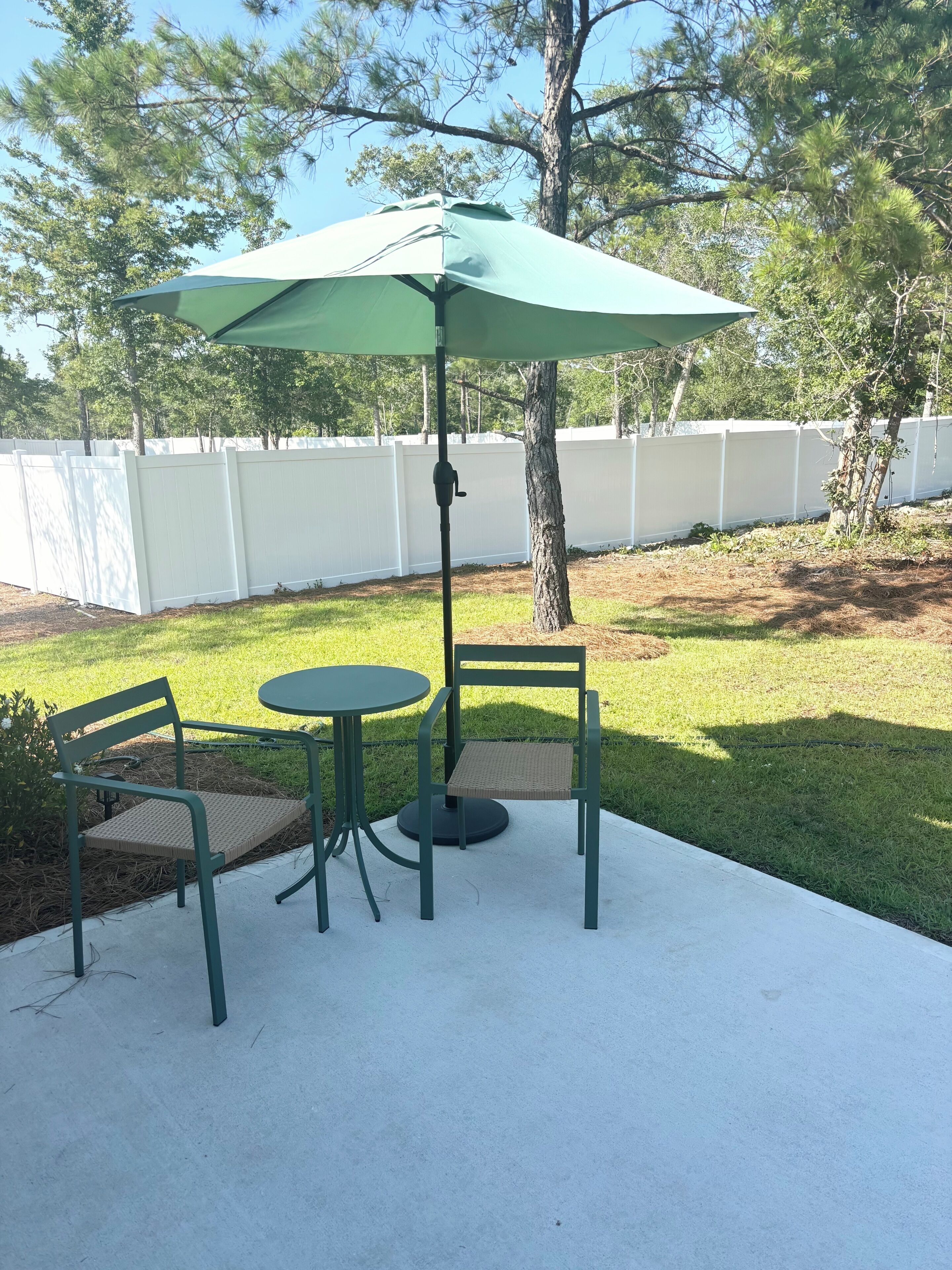 A patio with a table and two chairs, a green umbrella, and a white fence in the background.