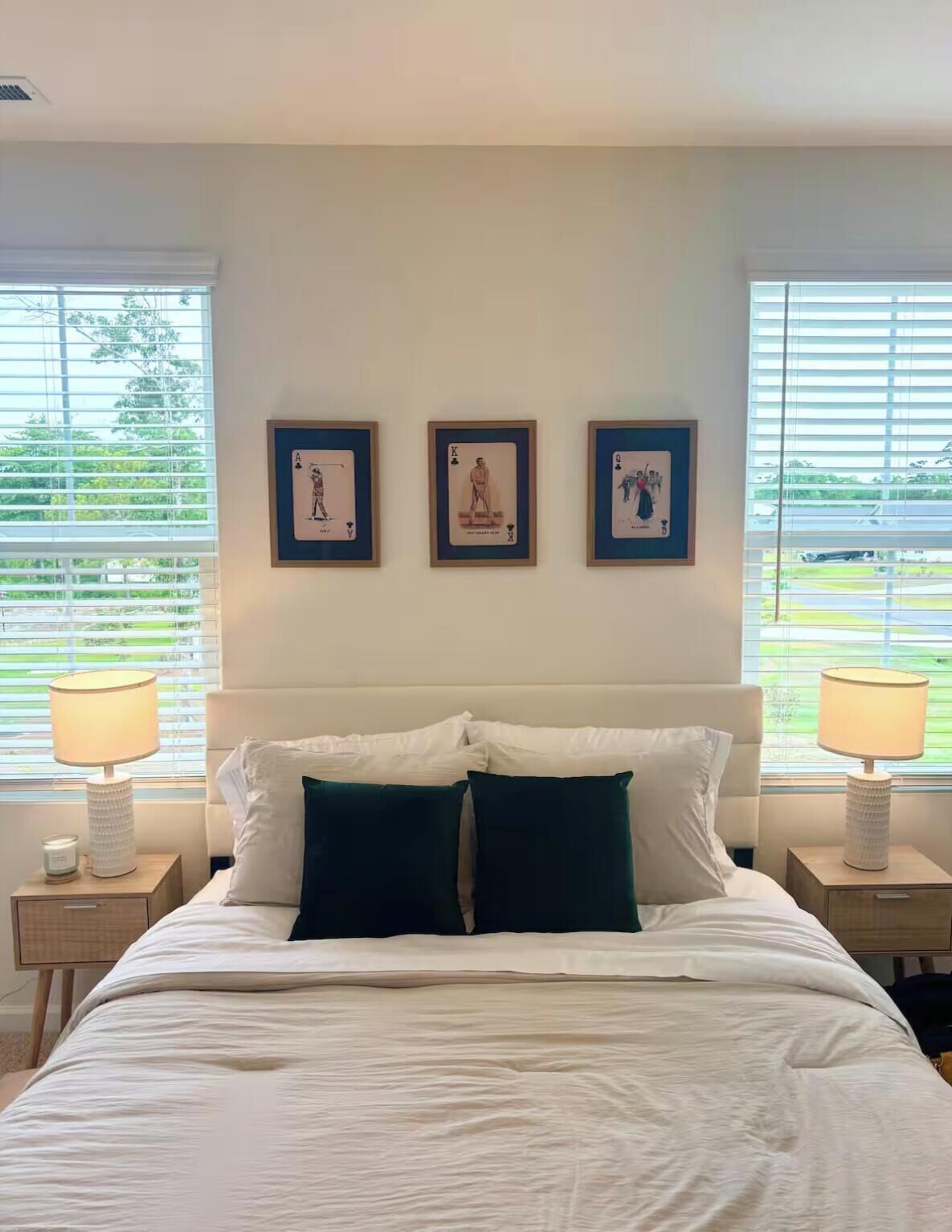 A neatly made bed with white and dark green pillows, flanked by two bedside lamps, and three framed artworks on the wall.