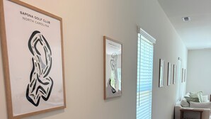 A hallway with framed artwork on the walls, a white couch, and a bookshelf. - Golf + Beach stay w/ private putting green! (Shallotte)