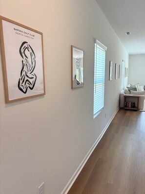 A hallway with framed artwork on the walls, a white couch, and a bookshelf. - Golf + Beach stay w/ private putting green! (Shallotte)
