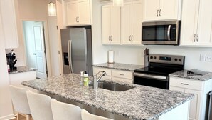 A modern kitchen with white cabinetry, a granite island with bar stools, stainless steel appliances, and pendant lighting. - Golf + Beach stay w/ private putting green! (Shallotte)