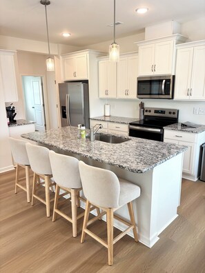 A modern kitchen with white cabinetry, a granite island with bar stools, stainless steel appliances, and pendant lighting. - Golf + Beach stay w/ private putting green! (Shallotte)