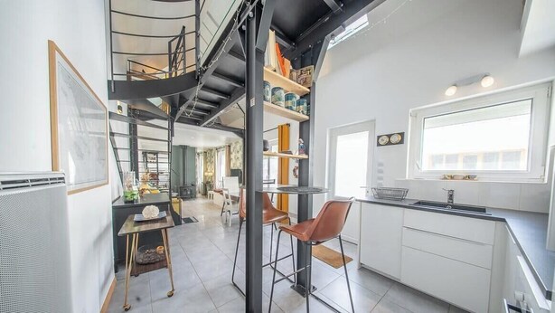 Una cocina moderna con estufa integrada, una zona de comedor con una mesa redonda y sillas, y una escalera que sube a un nivel superior.
