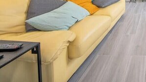 Una habitación con un sofá amarillo, un reposapiés marrón, una mesita con un control remoto y una pared blanca con una planta.