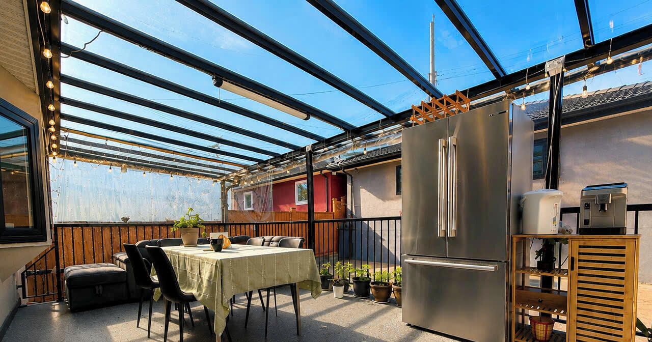 Un espacio de comedor al aire libre techado con una mesa, sillas, un refrigerador y plantas en macetas.