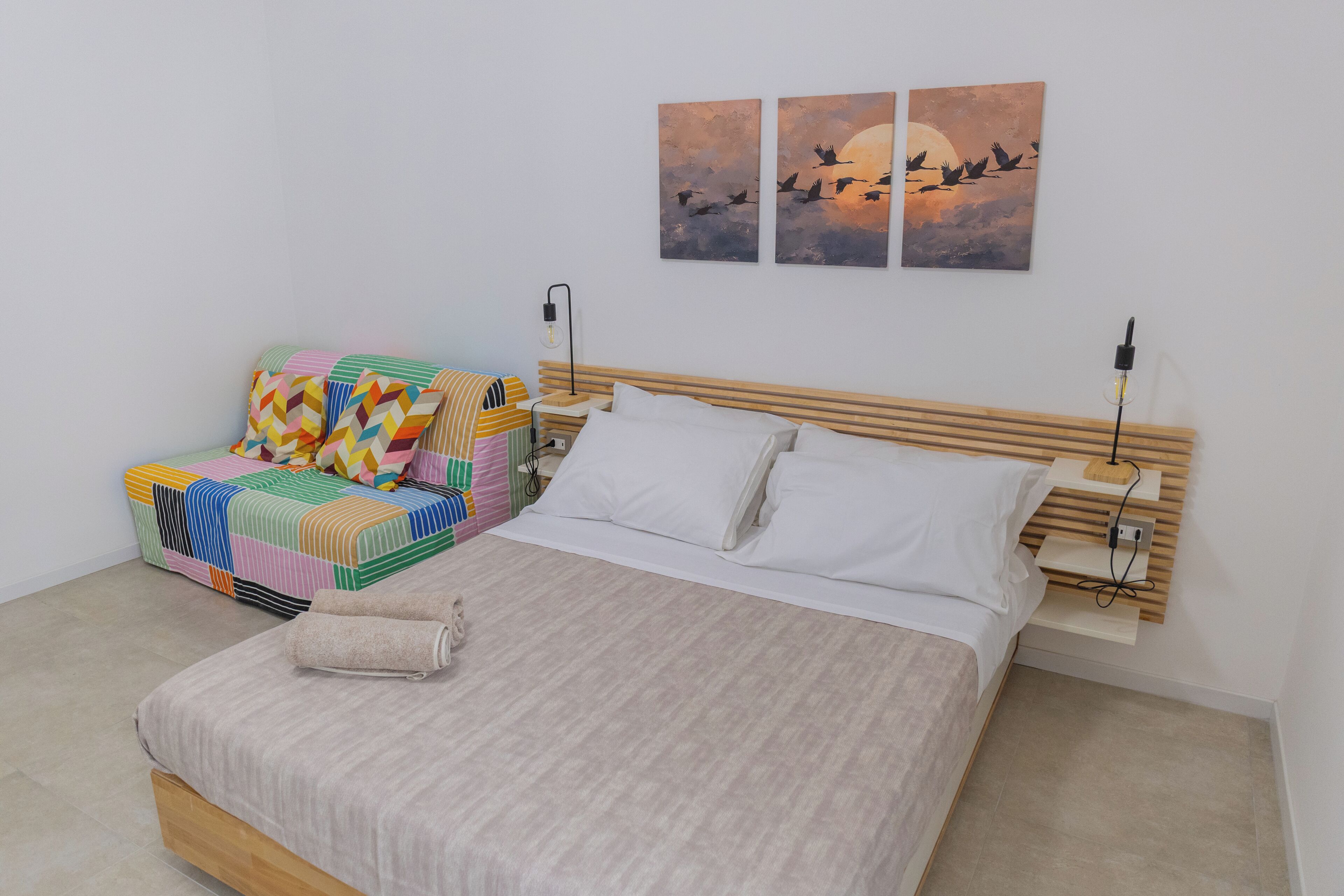 A bedroom with a bed, a colorful sofa, a nightstand, and wall art.