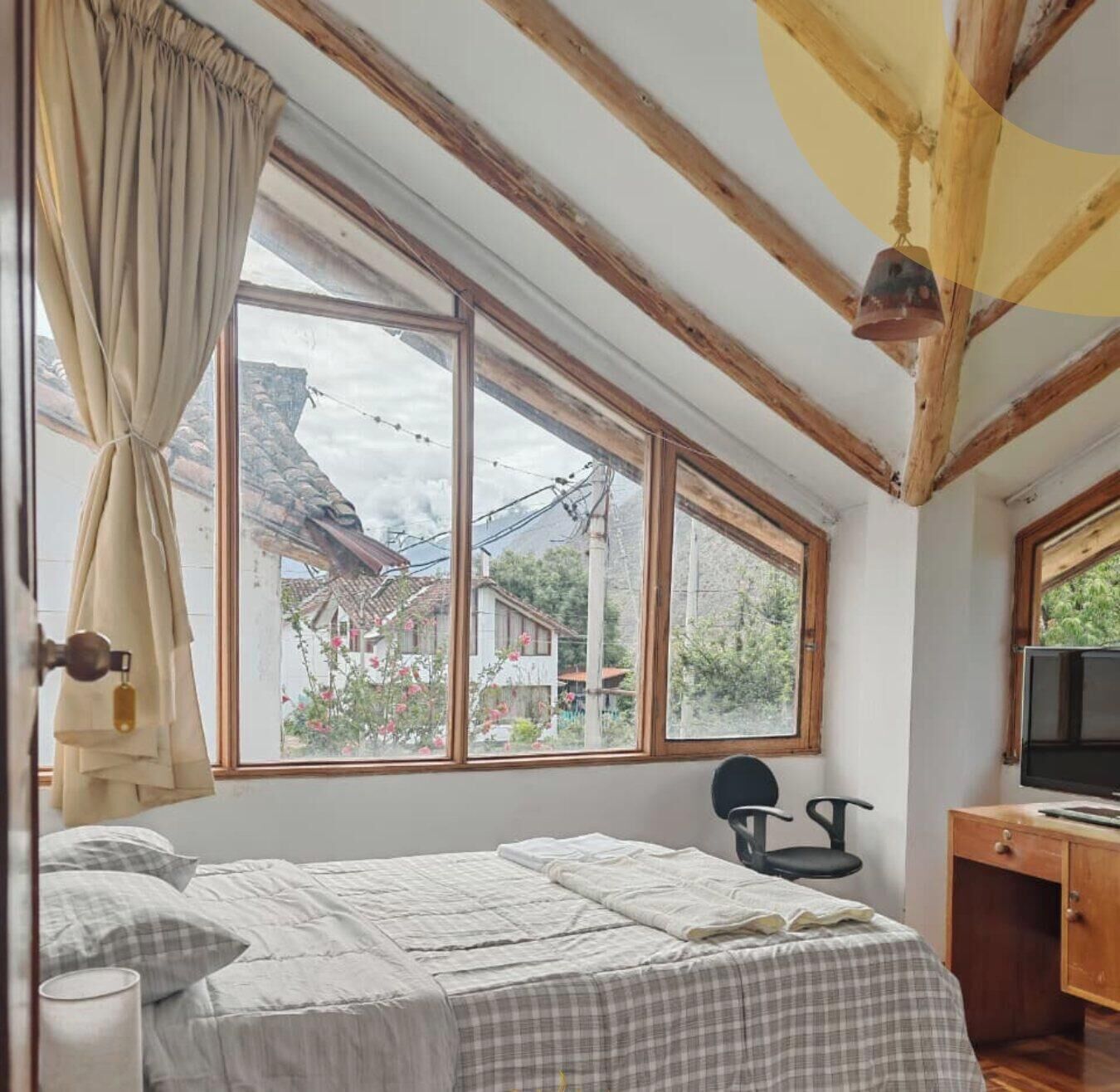 Une chambre avec une grande fenêtre, des poutres en bois, un lit, une chaise et un bureau avec une télévision.
