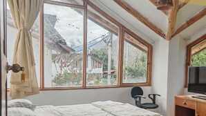 A bedroom with a large window, wooden beams, a bed, a chair, and a desk with a television. - Umay Lodge: A House in the Mountains (Urubamba)