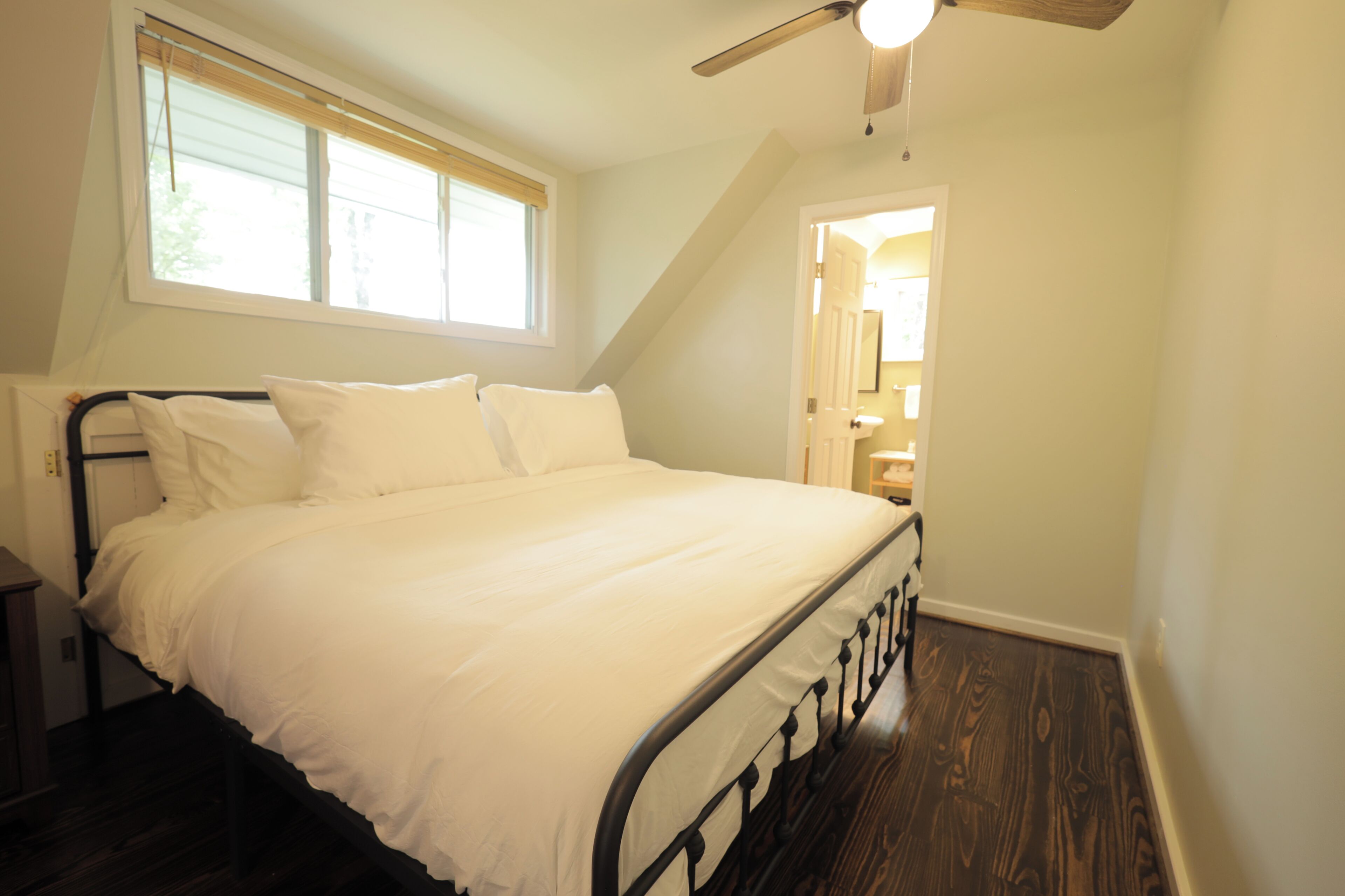 A bedroom with a bed, a window, a ceiling fan, and a door leading to another room.