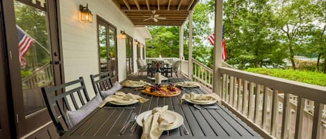 A covered outdoor dining area with a table set for two, chairs, and a view of the surrounding greenery.