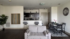Una sala de estar moderna con un sofá gris, un comedor con mesa y sillas, una cocina con microondas y horno, y un reloj grande en la pared.