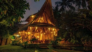 A wooden structure with a sloped roof, surrounded by lush greenery and palm trees. - Cacao House Eco-luxe Family Home Near Green School (Abiansemal)