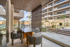 A modern outdoor dining area with a glass partition, wooden furniture, and a view of a multi-story building with balconies. - Peaceful Condo With Resort Pool, High-speed Wifi & Relaxing Outdoor Space (Cabo San Lucas)