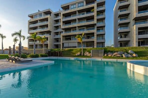 A modern hotel with a large swimming pool, multiple balconies, and palm trees. - Peaceful Condo With Resort Pool, High-speed Wifi & Relaxing Outdoor Space (Cabo San Lucas)