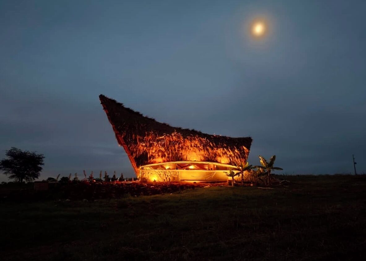 A thatched-roof building with a circular structure, illuminated by warm lighting, set against a night sky with a visible moon.