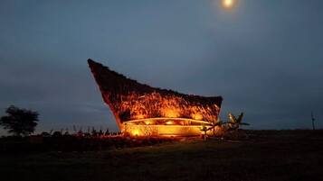 A thatched-roof building with a circular structure, illuminated by warm lighting, set against a night sky with a visible moon.