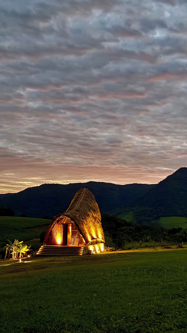 A thatched-roof structure with a staircase leading up to a door, surrounded by a grassy field and mountains in the background.