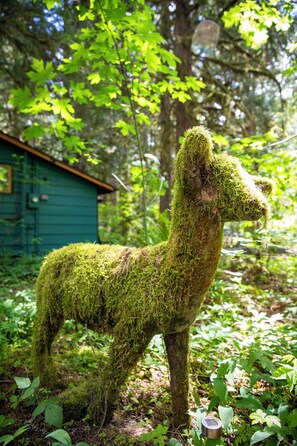 Eine moosbewachsene Statue eines Lamas in einer bewaldeten Gegend, im Hintergrund ein kleines Gebäude.