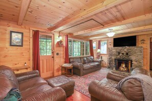 Ein gemütliches Blockhaus-Interieur mit Kamin aus Stein, Ledersofas, Holzwänden und einem großen Fenster.