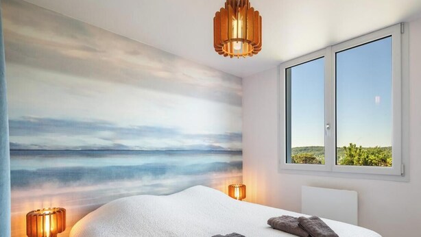 A bedroom with a large bed, two bedside lamps, a view of the sea through a window, and a ceiling light. - Gîtes de France® - La Villa Pomponette (CRIEL SUR MER)
