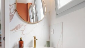 A bathroom with a marble wall, a round mirror, a white sink with gold fixtures, and a window above the mirror. - Gîtes de France® - La Villa Pomponette (CRIEL SUR MER)