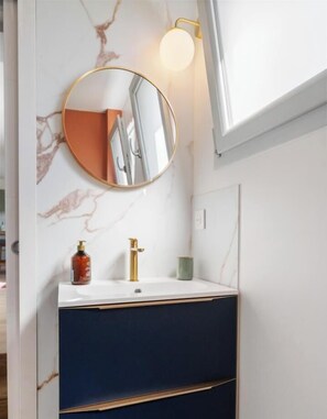 A bathroom with a marble wall, a round mirror, a white sink with gold fixtures, and a window above the mirror. - Gîtes de France® - La Villa Pomponette (CRIEL SUR MER)