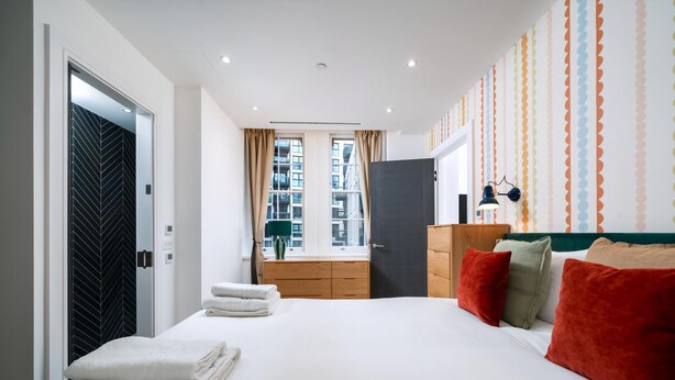 Ein Hotelzimmer mit einem Bett, einem Schminktisch, einem Nachttisch, einem Fenster mit Vorhängen und Blick auf Gebäude.
