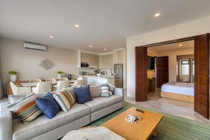 A modern living room with a sofa, coffee table, and dining area. There is a kitchen in the background with a refrigerator and microwave. - Peaceful Coastal Stay With Pool, Balcony & Fitness Center (Cabo San Lucas)
