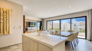 Cozinha moderna com ilha central, área de refeições com cadeiras, televisão e uma janela grande com vista para a cidade.