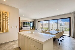 A modern kitchen with a central island, dining area with chairs, a TV, and a large window with a city view. - Peaceful Coastal Stay With Pool, Balcony & Fitness Center (Cabo San Lucas)