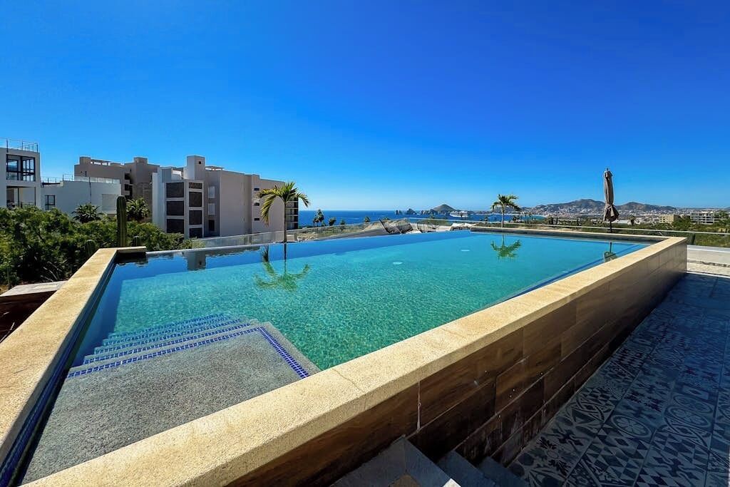 Ein rechteckiger Pool mit klarem Wasser, umgeben von einer gefliesten Terrasse, mit Gebäuden und blauem Himmel im Hintergrund.