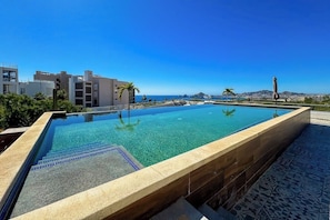 A rectangular pool with clear water, surrounded by a tiled deck, with buildings and a clear sky in the background. - Peaceful Coastal Stay With Pool, Balcony & Fitness Center (Cabo San Lucas)