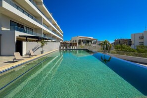 A clear swimming pool with lounge chairs, a modern building with balconies, and a clear blue sky. - Peaceful Coastal Stay With Pool, Balcony & Fitness Center (Cabo San Lucas)