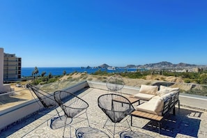 A rooftop terrace with outdoor furniture, a clear view of the ocean, and distant mountains. - Peaceful Coastal Stay With Pool, Balcony & Fitness Center (Cabo San Lucas)