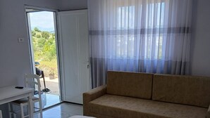 Une pièce comprenant un lit, un canapé, une table et une chaise. Il y a une fenêtre avec des rideaux, et de là, on peut admirer la verdure à l’extérieur.