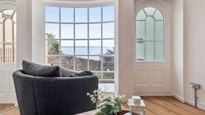 Ein Zimmer mit einer gewölbten Erkerfensterfront, einem dunkelgrauen Sofa, einem kleinen Tisch mit einer Pflanze und einer weißen Tür mit einer Milchglasverkleidung.