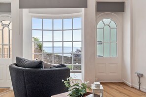 A room with a curved bay window, a dark grey sofa, a small table with a plant, and a white door with a frosted glass panel. - Tenby Harbour View - Heart of Tenby, Sea Views (Tenby)