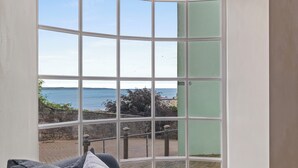 Ein Zimmer mit einem großen Erkerfenster mit Blick auf eine Küstenlandschaft, einer Steinmauer und einem gepflasterten Weg.