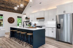 Eine moderne Küche mit weißen Schränken, einem Edelstahlkühlschrank, einer großen Kochinsel mit Sitzgelegenheiten und einer Holzbalkendecke.