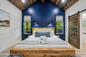 Ein Schlafzimmer mit einem Zargenholzboden, einem hölzernen Kopfteil, einem Bett mit weißen Leinen, zwei Nachttischlampen und Blick ins Grüne durch die Fenster.