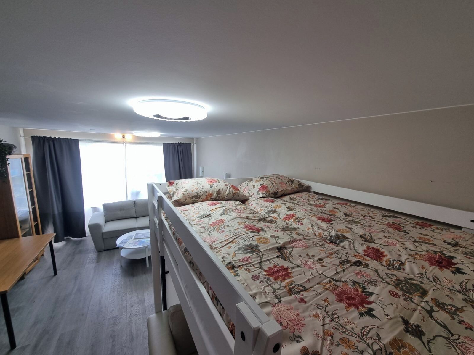 Ein kleiner, gut beleuchteter Raum mit einem Bett, einer Couch, einem Tisch und einem Fenster mit Vorhängen.