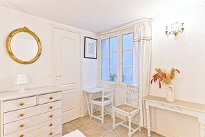 Ein Zimmer mit einem weißen Schminktisch, einem Sessel, einem Schreibtisch, einem Fenster mit Vorhängen, einer Chandelüster und einem gerahmten Bild an der Wand.